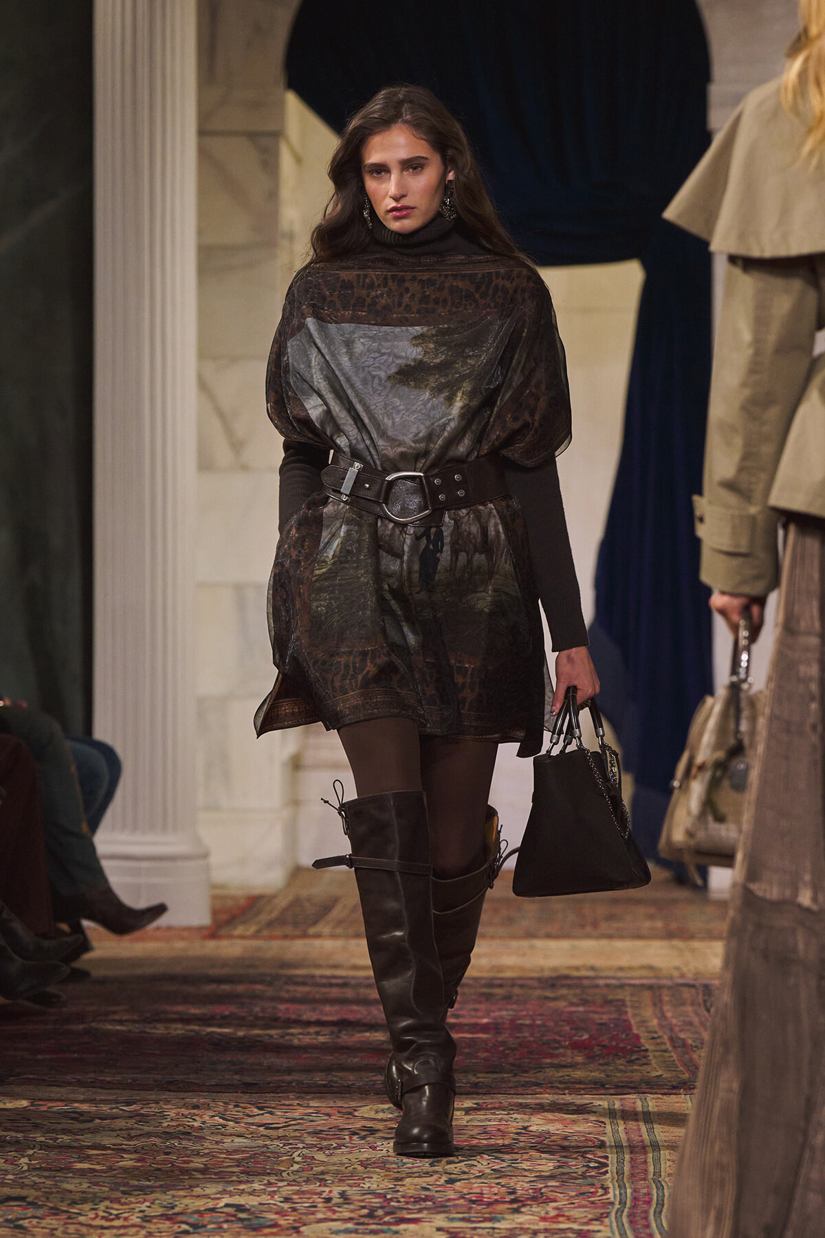 Model walking a runway in a dark, patterned cape-style dress cinched with a wide belt, paired with tall black boots and a black handbag.