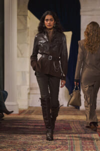 Model walking a runway in a dark brown leather belted jacket with chain details and knee-high boots.