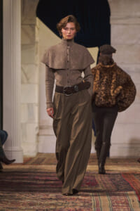Runway model wearing a textured taupe capelet over a high-neck knit top with wide-leg brown trousers and a thick black belt; leopard-print coat seen on a second model in the background.