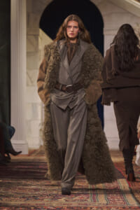 Model walks a runway in a gray suit with a long brown shearling coat and wide belt, on a patterned rug.