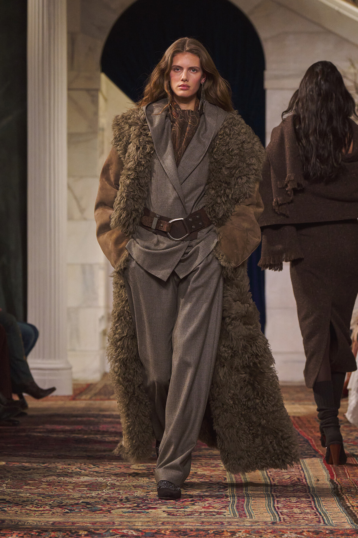 Model walks a runway in a gray suit with a long brown shearling coat and wide belt, on a patterned rug.