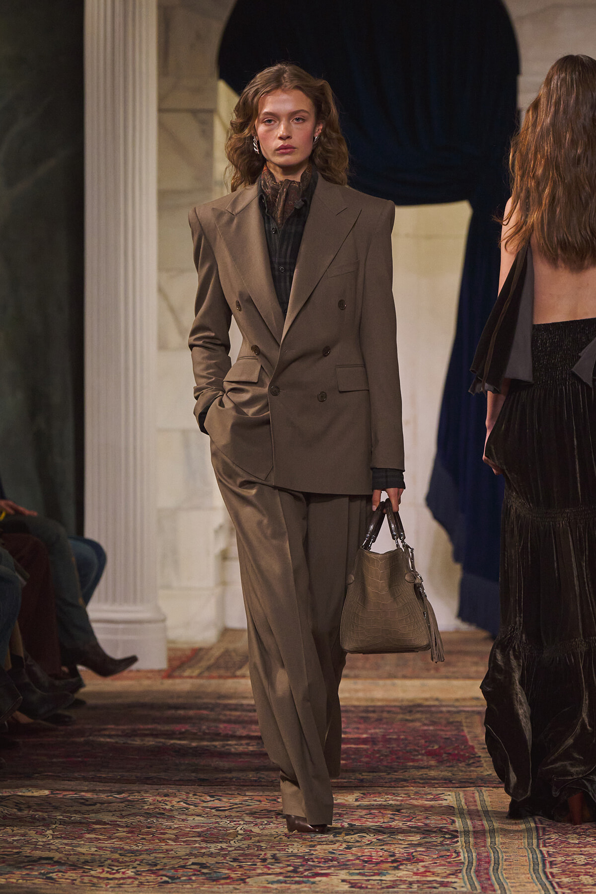 Model walking a runway in a taupe double-breasted suit with wide-leg pants, holding a beige crocodile-textured handbag.