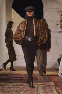 Model walks the runway in a leopard-print faux fur jacket, black lace turtleneck, black pants, and a brown newsboy cap.