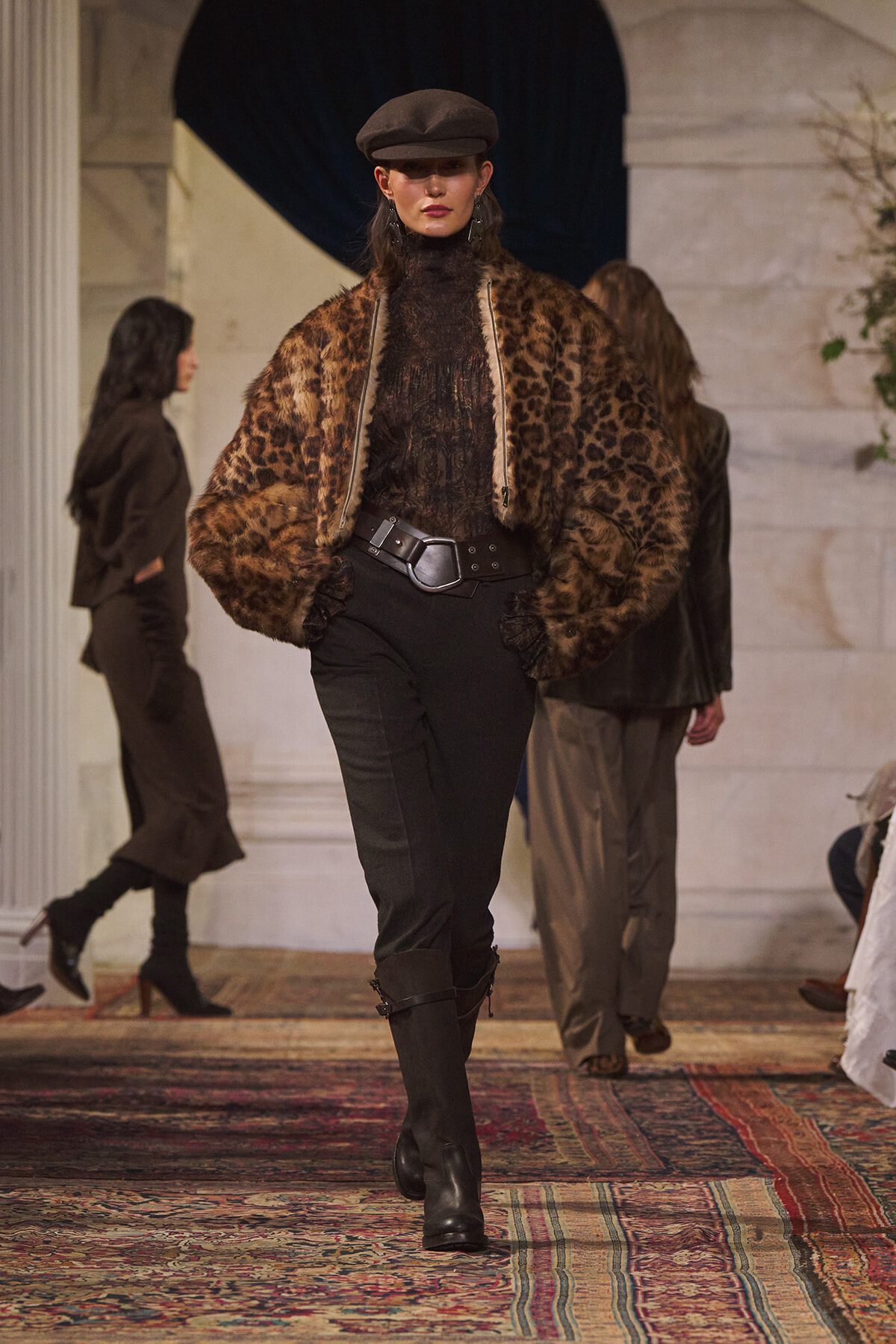 Model walks the runway in a leopard-print faux fur jacket, black lace turtleneck, black pants, and a brown newsboy cap.