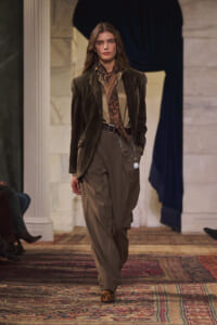 Model walking runway in brown velvet blazer, tan blouse, leopard scarf, and wide taupe trousers with chain accessory and leopard-print shoes