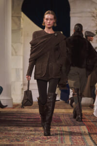 Model walks a runway in a dark brown textured wrap coat with a brooch, black ribbed tights, and tall black boots, carrying a small black handbag into the scene.
