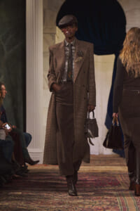Model on runway in a brown plaid overcoat, metallic shirt and tie, beret, and gray handbag.