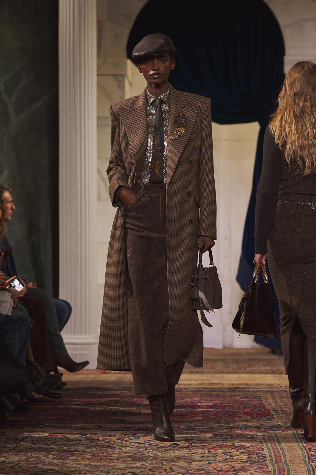 Model on runway in a brown plaid overcoat, metallic shirt and tie, beret, and gray handbag.