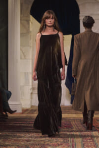 Model walking the runway in a long black velvet dress with thin straps and statement earrings; backstage-like architectural backdrop and patterned carpet visible.