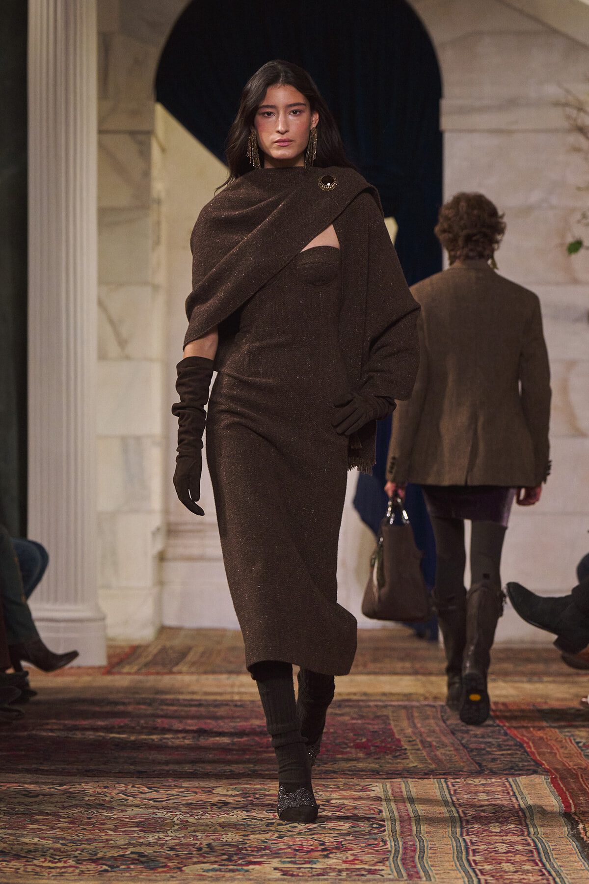 Model walks a runway in a brown shimmering knit ensemble: one-shoulder cape over a fitted dress, long brown gloves, and sparkly earrings in a marble-lined venue.