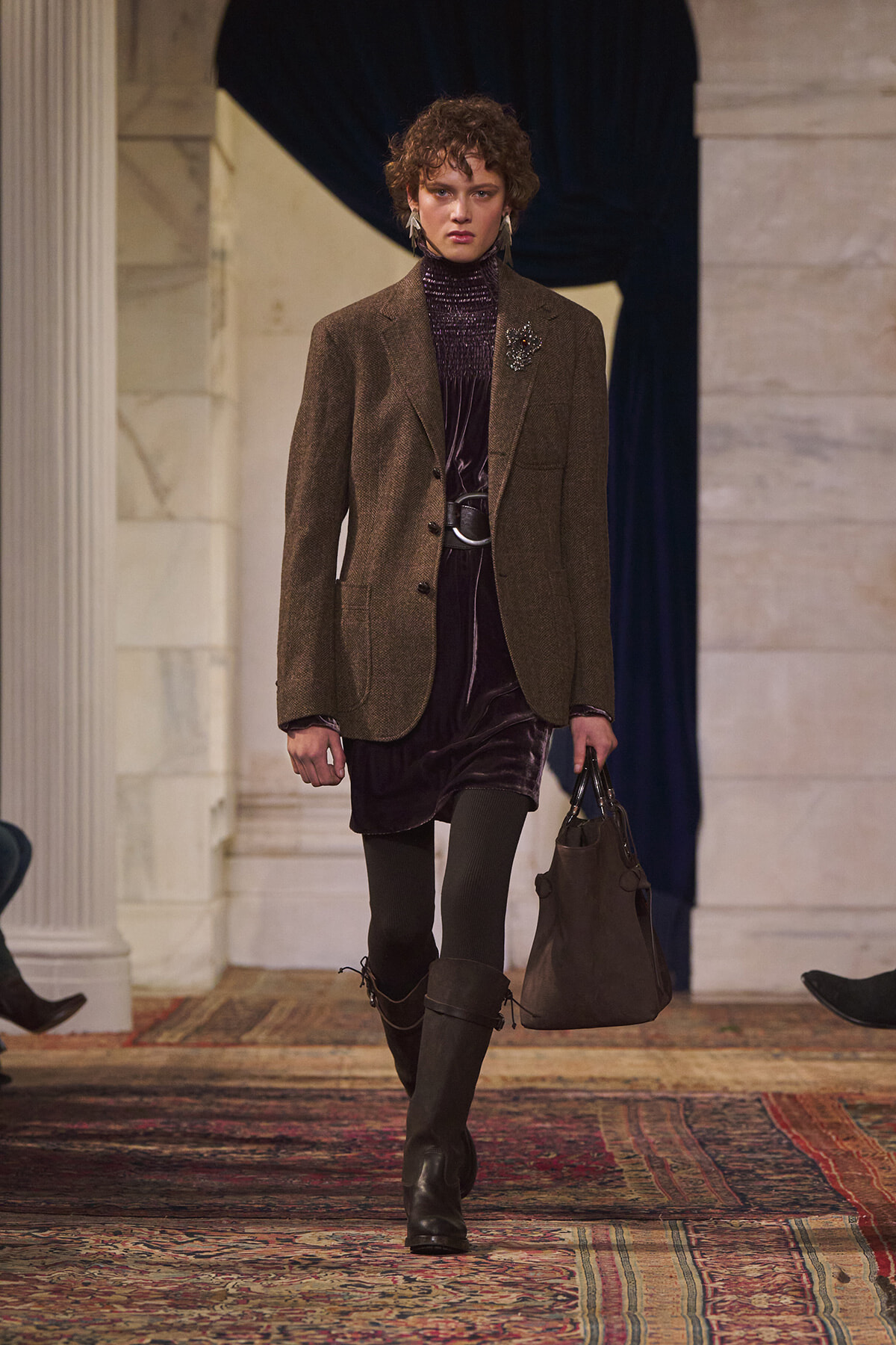 Model walking a runway in a brown blazer over a dark velvet dress, with tall boots and a brown handbag.