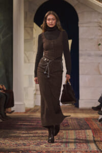 Model on a runway wearing a brown glittery sleeveless dress over a black turtleneck, with a silver chain belt and dark boots, carrying a brown handbag.