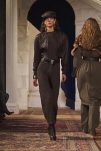 Model walking a runway in a black leather jacket over a dark knit jumpsuit with a wide belt and ankle boots.