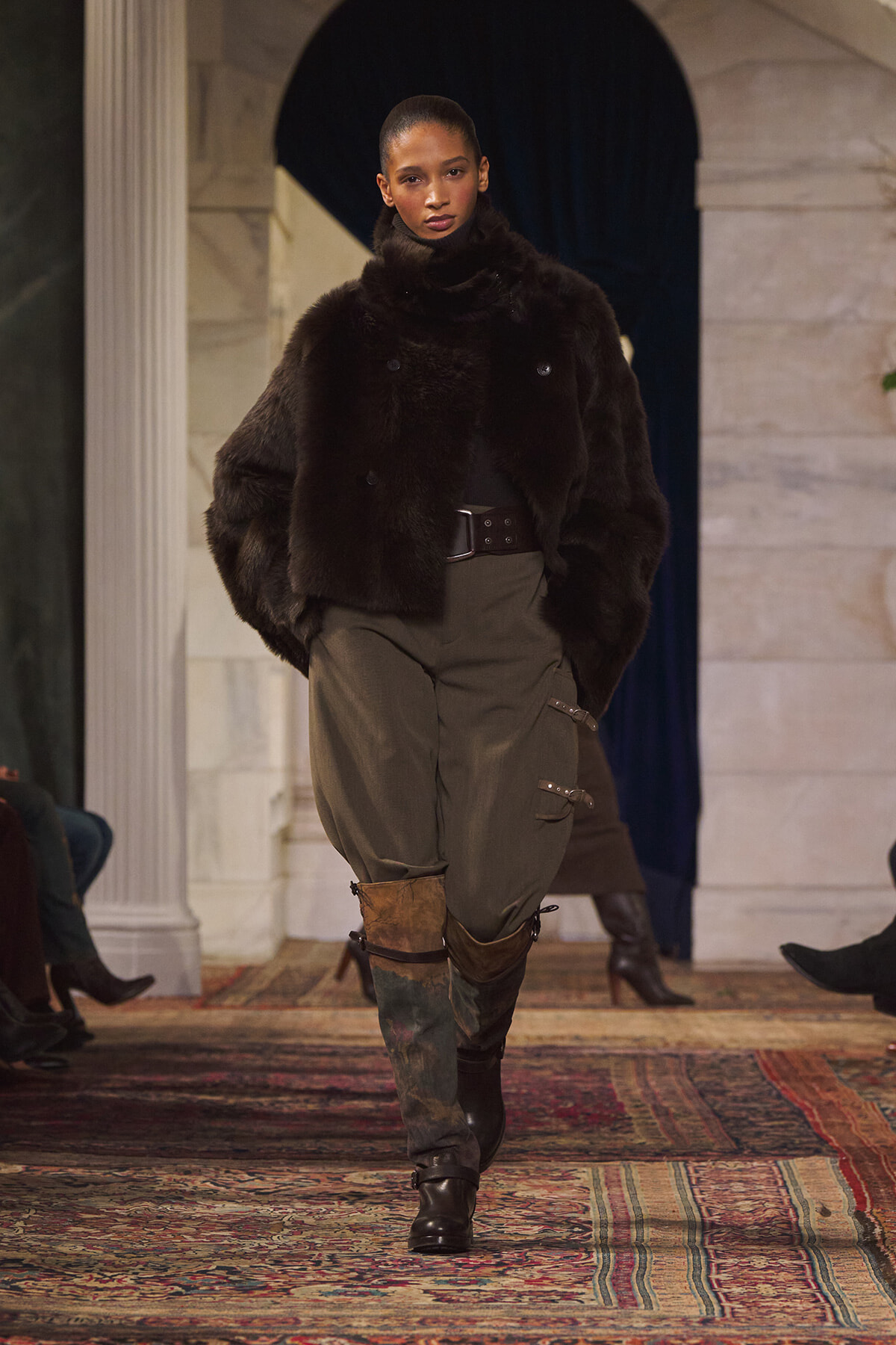 Model walks the runway in a black fur coat, olive cargo pants, and brown boots.