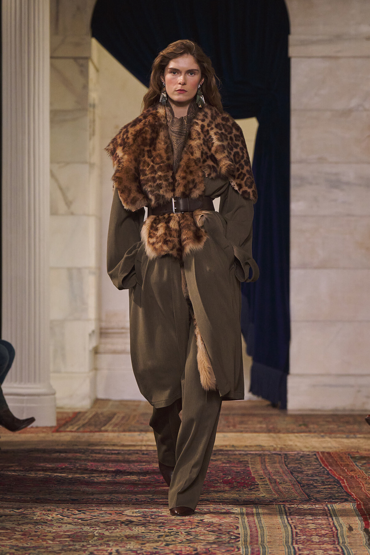 Model walking a runway in an olive-green suit with a leopard fur stole and wide belt.