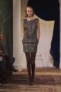 Model walks a runway in a metallic textured silver-gray sleeveless dress with pockets, wearing chandelier earrings.
