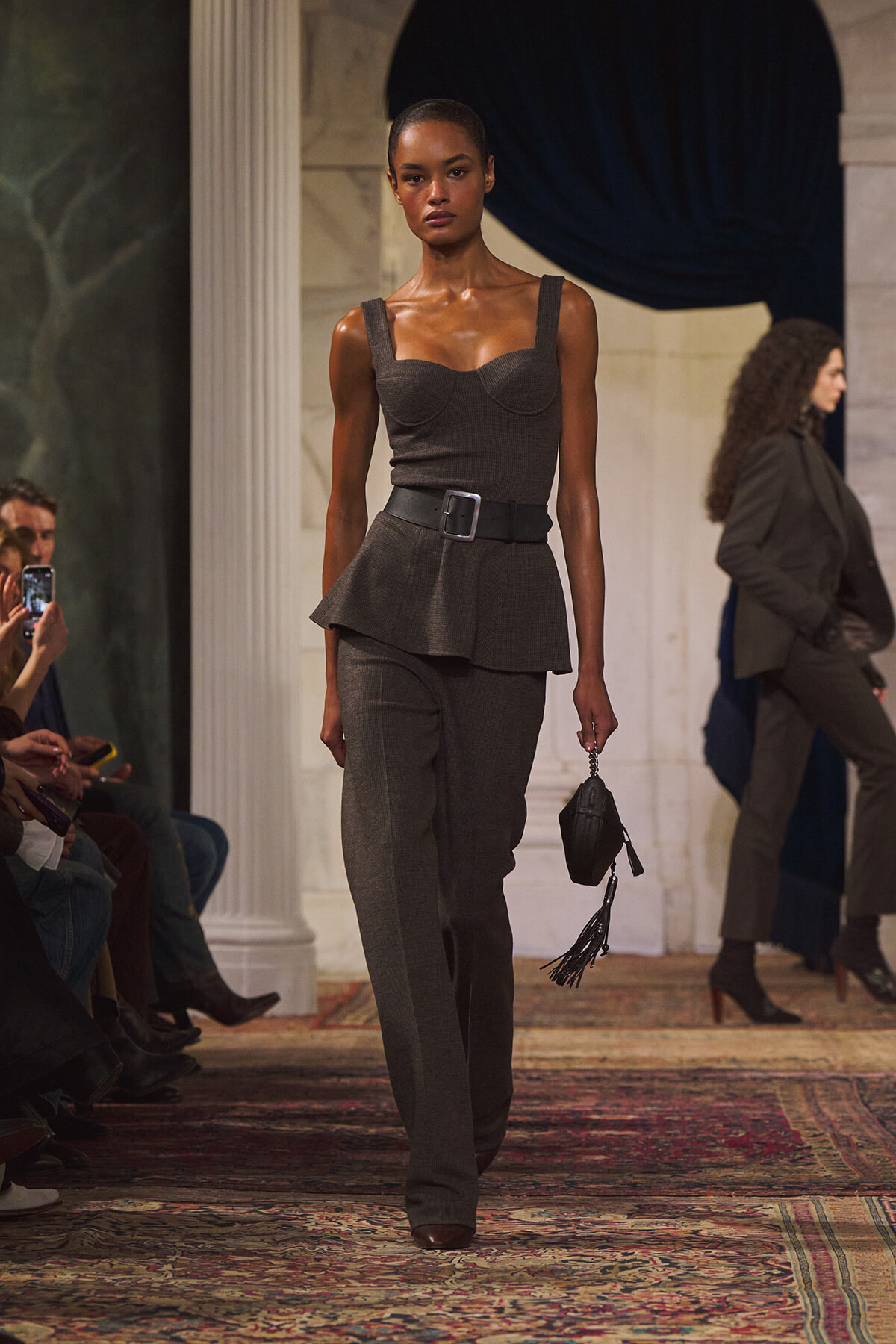 Model on a runway wearing a dark gray peplum top with wide straps, matching gray pants, a black belt, and a small black handbag.