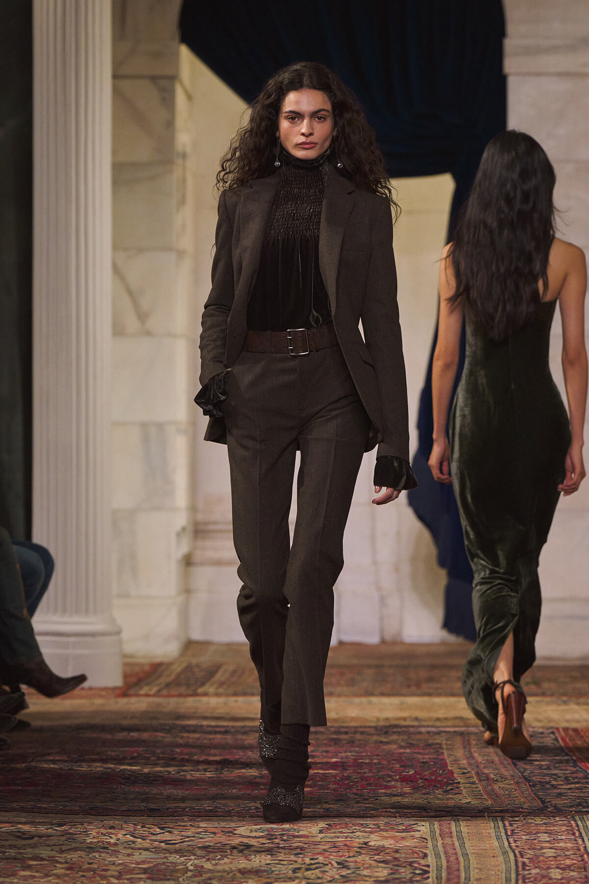 Model walks the runway in a charcoal tailored suit with blazer, matching pants, and a dark turtleneck; belt at the waist.