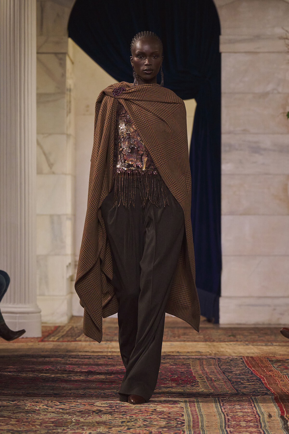 Fashion model walks the runway in a brown plaid cape draped over a sequined top and wide black trousers.