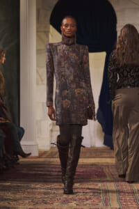 Model walking a runway in a metallic floral mini dress and tall black boots on a patterned carpeted floor.