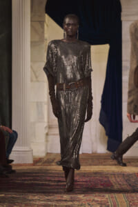 Model on a runway wearing a metallic silver sequin dress with long brown gloves and a wide brown belt in a marble venue.