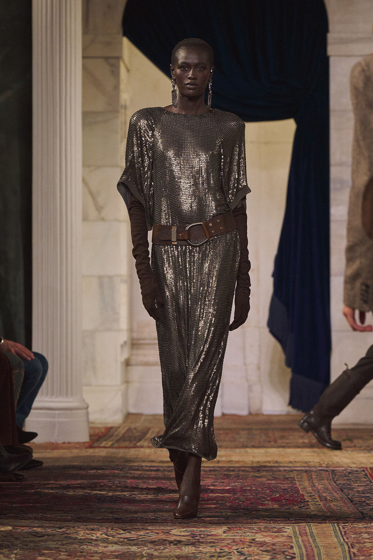 Model on a runway wearing a metallic silver sequin dress with long brown gloves and a wide brown belt in a marble venue.
