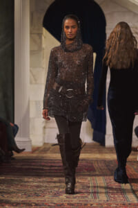 Model walking a runway in a black, sequined hooded top, with a wide belt, black leggings, and tall boots.