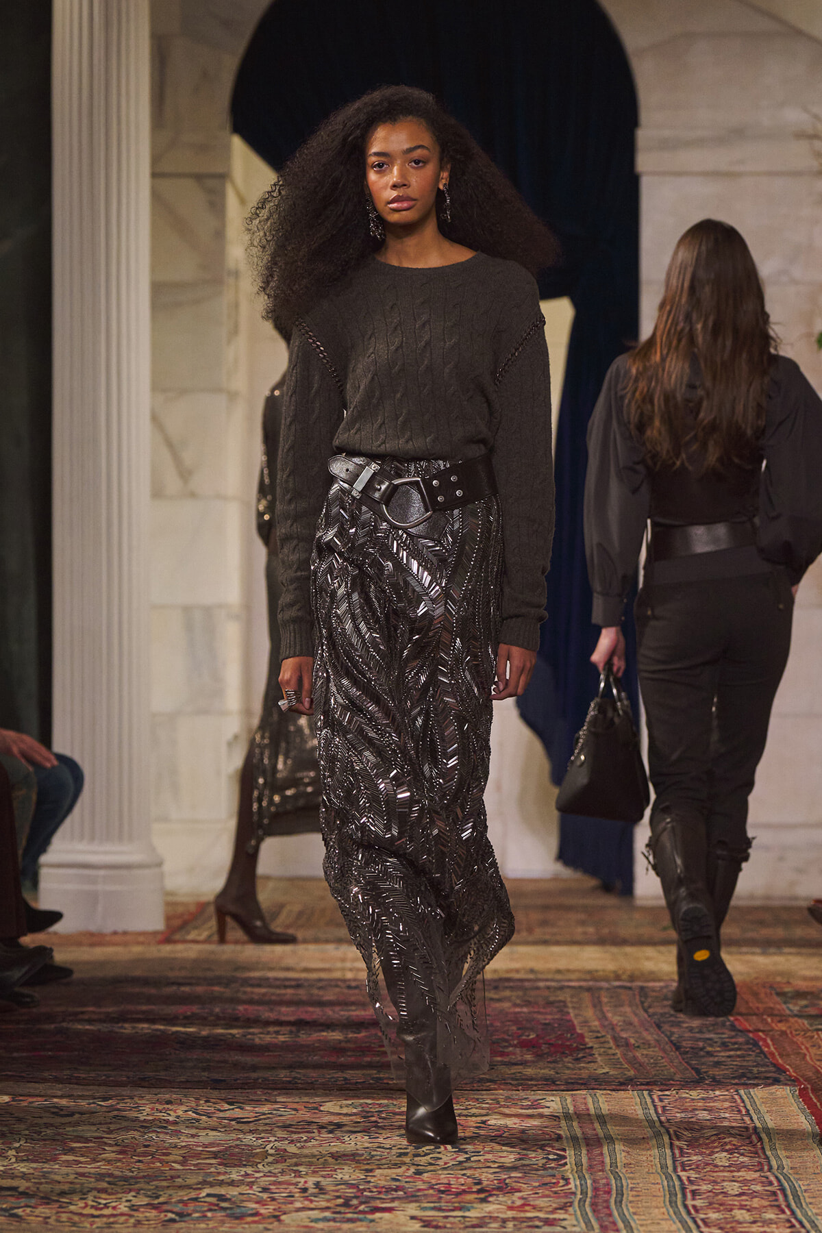 Model walks a runway wearing a dark knit sweater and a metallic, patterned midi skirt with a wide black belt at the waist.