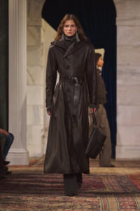 Model walking a runway in a long black leather trench coat with a belt, gray turtleneck, and matching gloves while carrying a black bag.