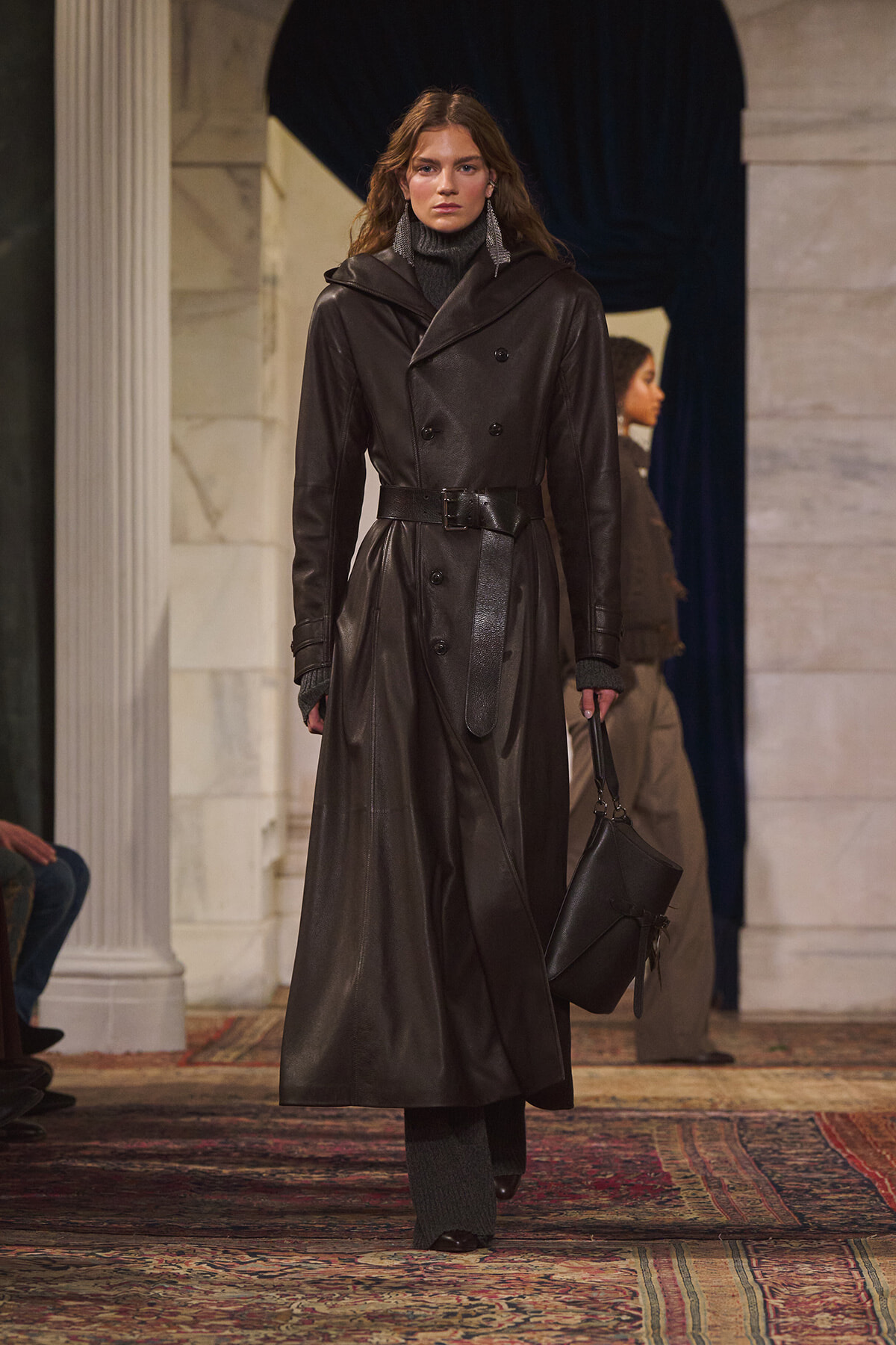 Model walking a runway in a long black leather trench coat with a belt, gray turtleneck, and matching gloves while carrying a black bag.