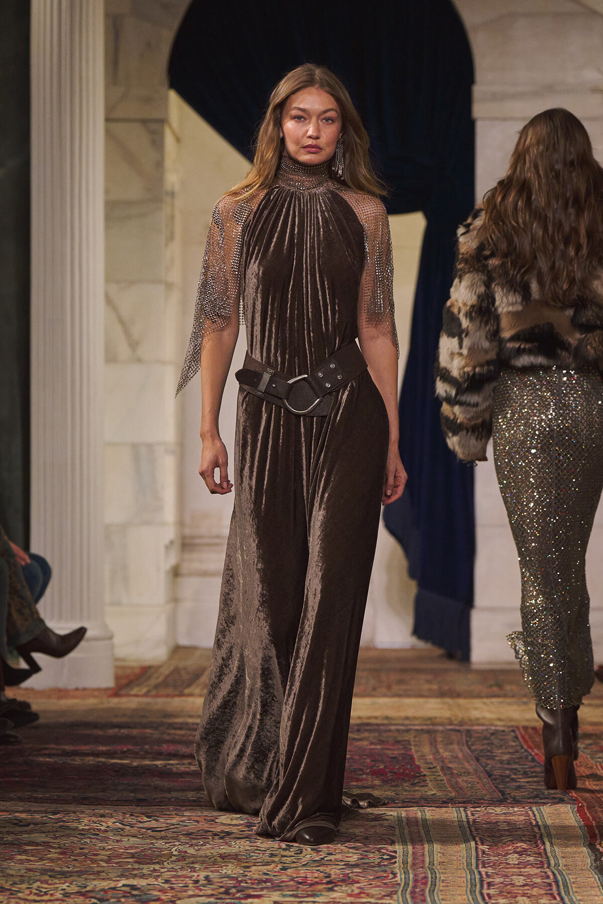 Model on a runway wearing a brown velvet jumpsuit with sheer, beaded sleeves and a wide black belt.