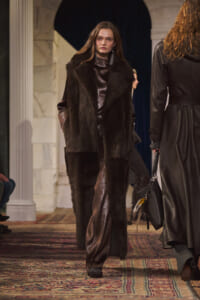 Model on a runway wearing a long dark fur coat over a metallic brown gown, with statement earrings, walking toward the camera in a venue with columns and a patterned carpet.