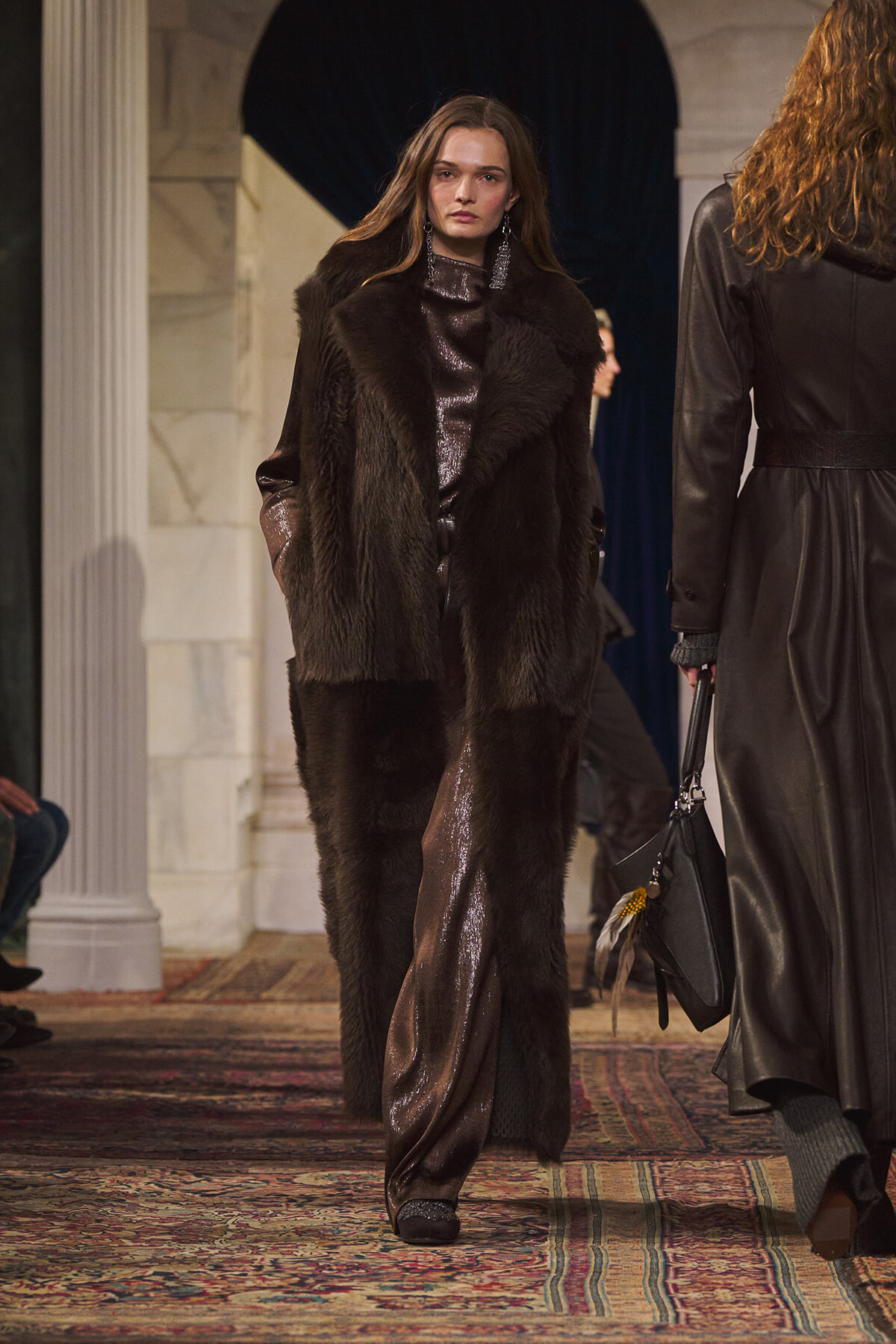 Model on a runway wearing a long dark fur coat over a metallic brown gown, with statement earrings, walking toward the camera in a venue with columns and a patterned carpet.