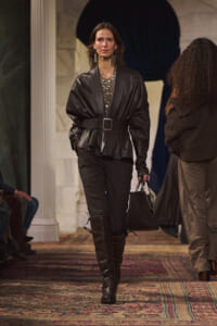 Model on a runway wearing a black leather belted peplum top, black pants, knee-high boots, and a black handbag.