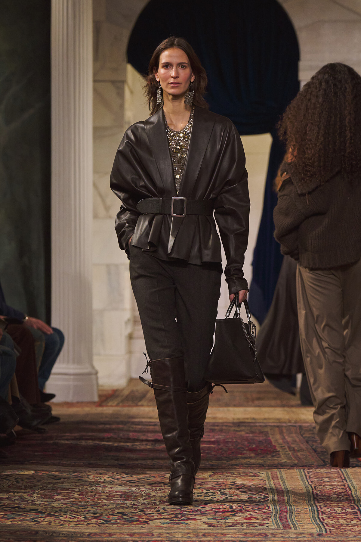 Model on a runway wearing a black leather belted peplum top, black pants, knee-high boots, and a black handbag.