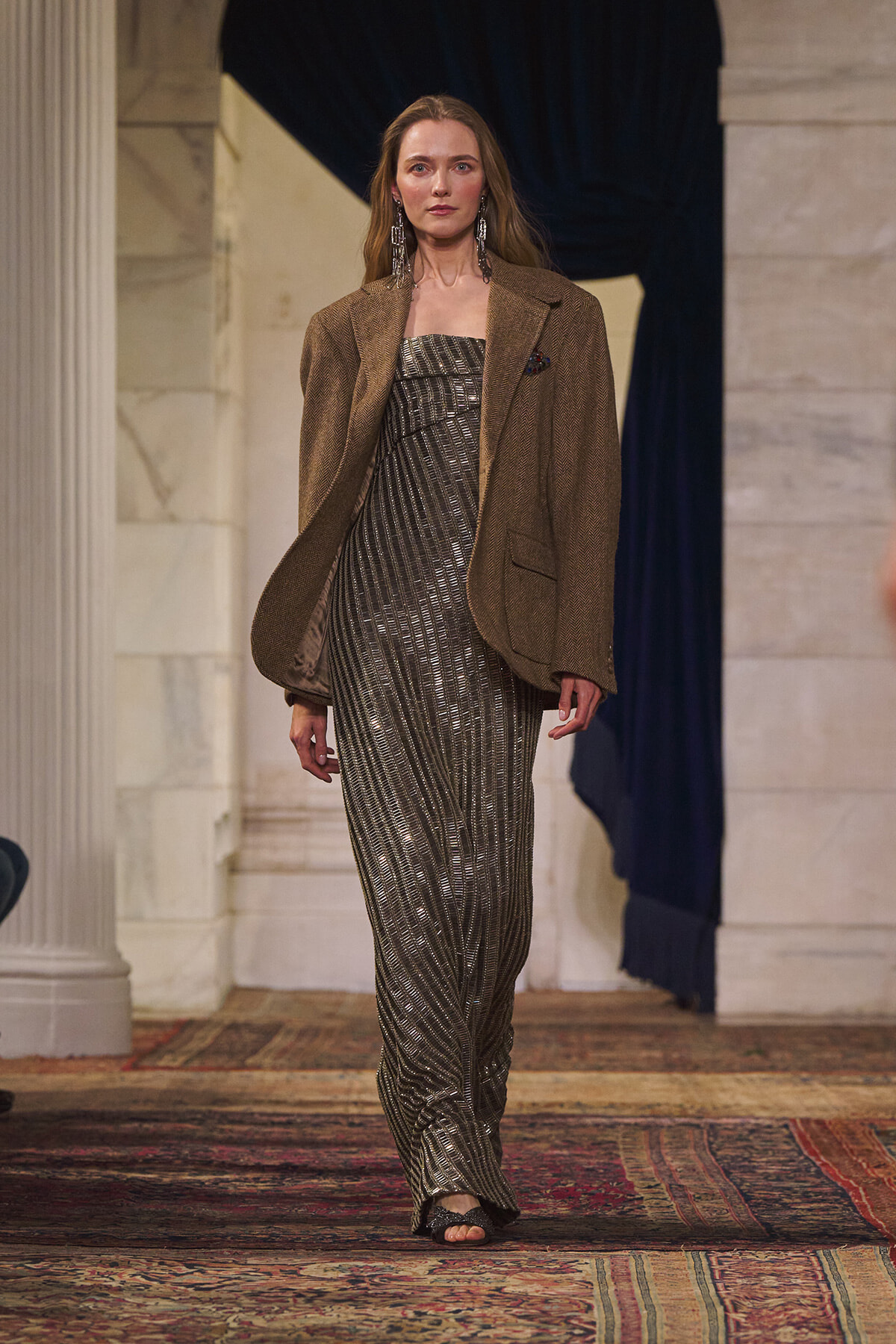 Model on a fashion runway wearing a metallic silver-striped strapless jumpsuit with a brown oversized blazer and statement earrings, walking the catwalk in a grand venue.