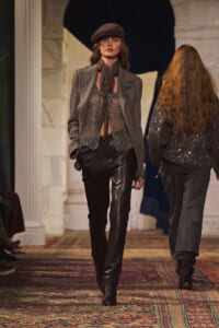 Model walking a fashion runway in a textured blazer, chainmail top, scarf, leather pants, and a brown beret.