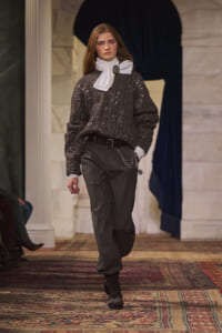Model walking a runway in a dark metallic knit sweater, white neck scarf with brooch, gray trousers, belt and chain accessory.