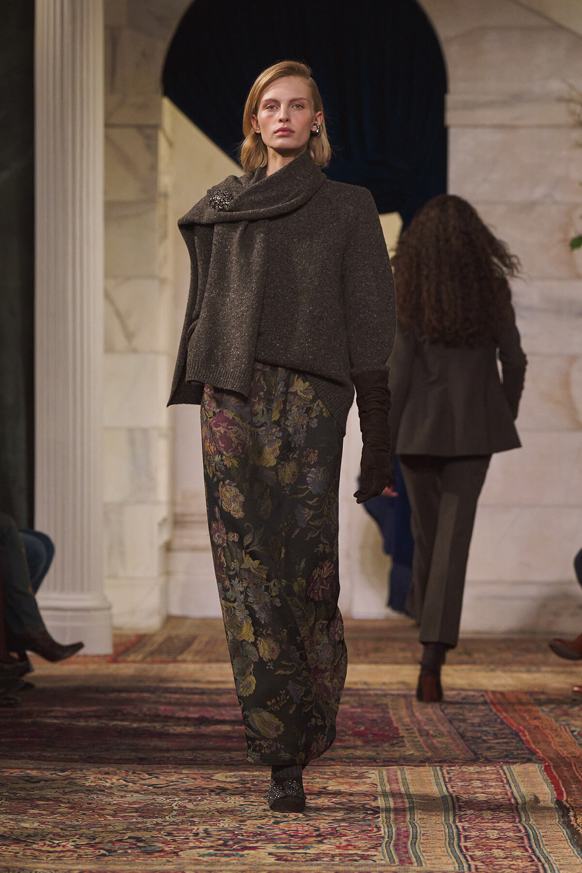 Model walking a runway in a dark, shimmery cape-style sweater with scarf collar and floral print wide-leg pants, wearing gloves, on a patterned carpeted catwalk.
