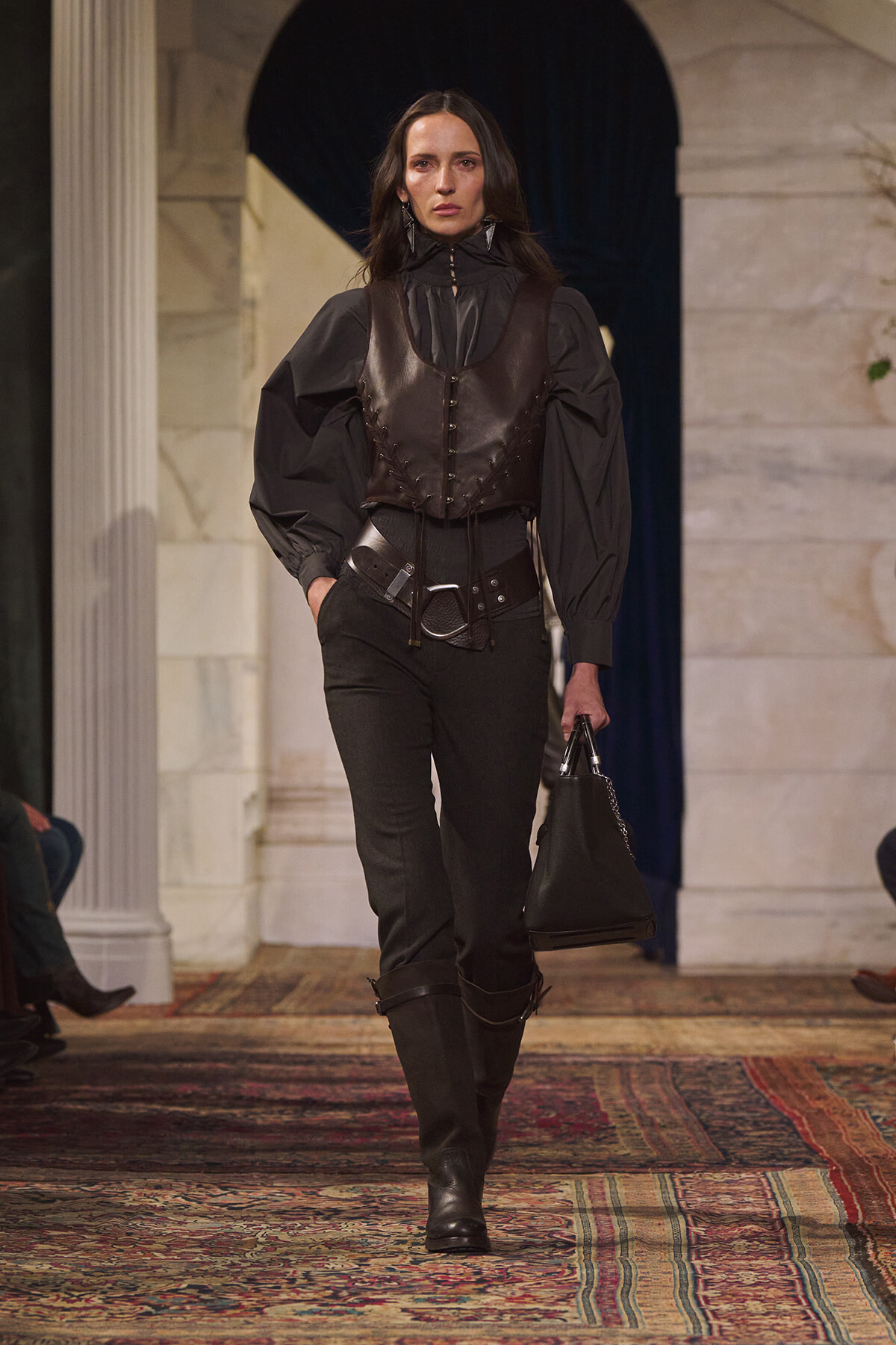 Model on a fashion runway wearing a dark ensemble: brown leather corset vest over a black high-neck blouse with voluminous sleeves, black pants, knee-high boots, and a black handbag.