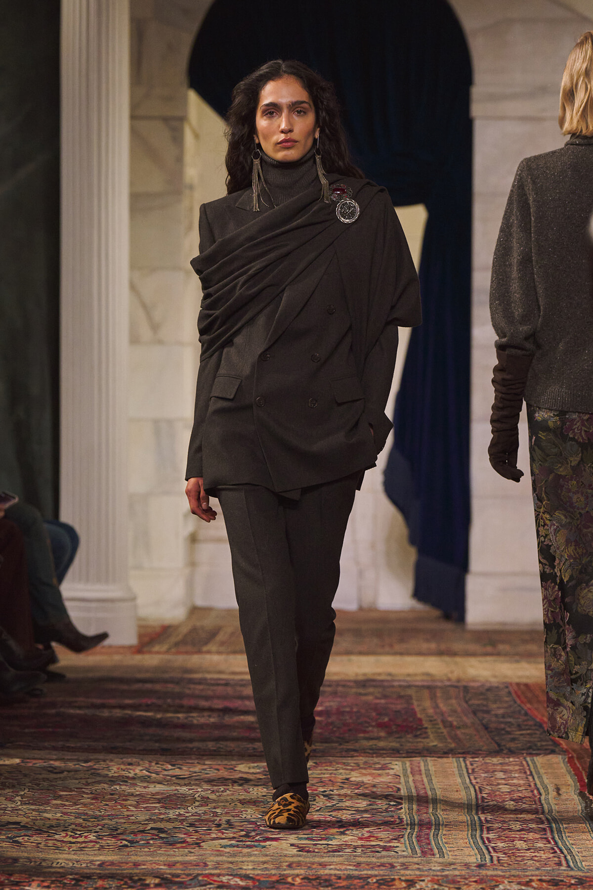 Model on a runway wearing a dark draped blazer with a scarf-like overlay, black pants, and leopard-print shoes.