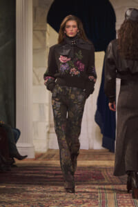 Runway model wearing a dark floral knit sweater with a large black bow and matching floral trousers, walking the catwalk.
