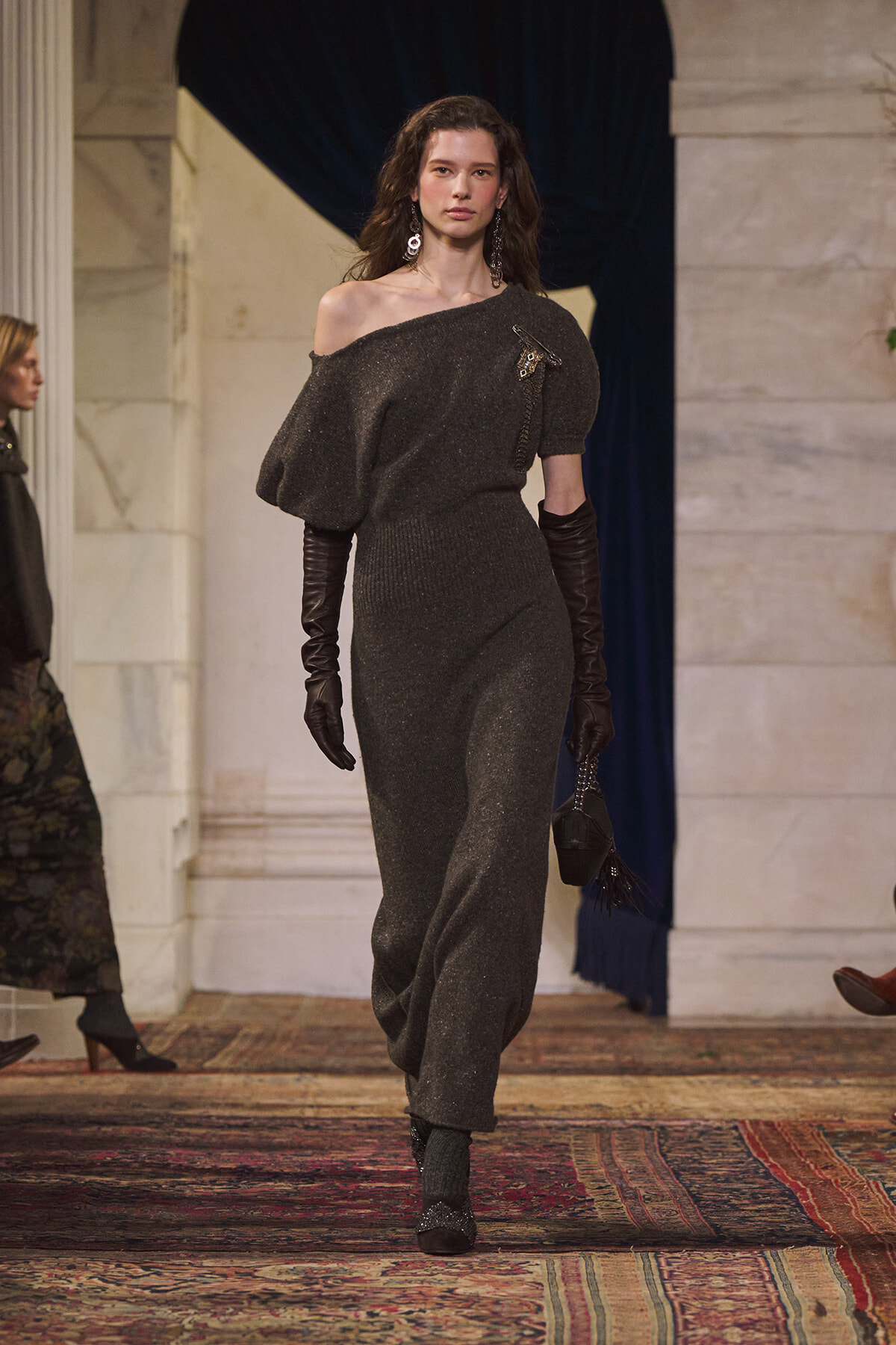 Model walking a runway in a sparkly dark gray off-shoulder jumpsuit with a voluminous one-shoulder sleeve, black opera gloves, dangling earrings, and a small black handbag.