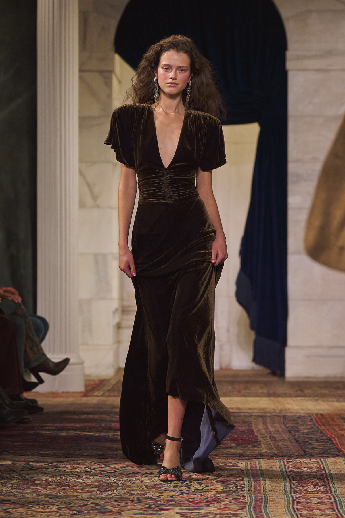 Model walking a runway in a dark velvet gown with a deep V neckline and ruched waist, clutching the skirt as she steps forward.