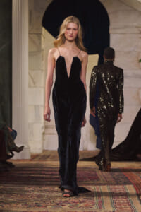 Model walks the runway in a floor-length navy velvet gown with a plunging V-neckline; another sequined outfit is in the background.