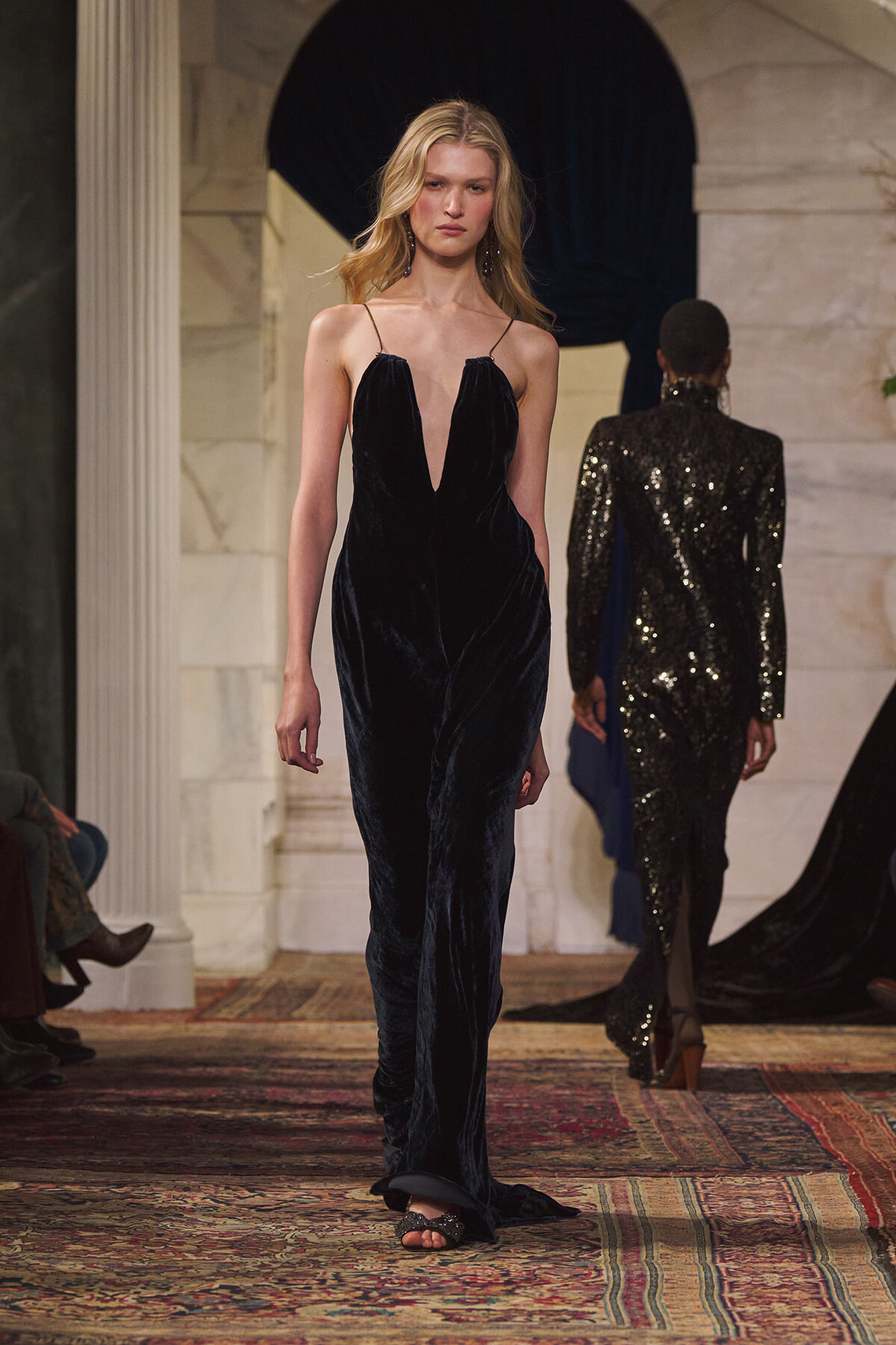 Model walks the runway in a floor-length navy velvet gown with a plunging V-neckline; another sequined outfit is in the background.