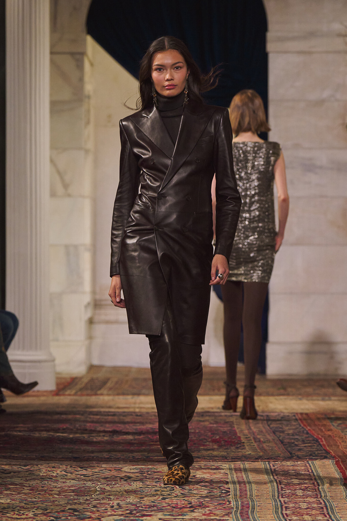 Model walking the runway in a black leather double-breasted blazer dress with a turtleneck and knee-high boots, leopard-print shoes showing at the toes.