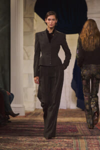 Female model walks a runway in a dark tailored suit with a fitted blazer and wide-leg trousers, in a marble-columned venue.
