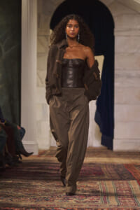 Model walking a runway in a brown leather corset, oversized knit cardigan, and taupe tailored trousers under warm venue lighting.