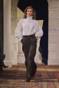 Model walking runway in a white high-neck blouse with voluminous sleeves and black wide-leg trousers, accessorized with a belt and layered chains in an elegant interior setting.